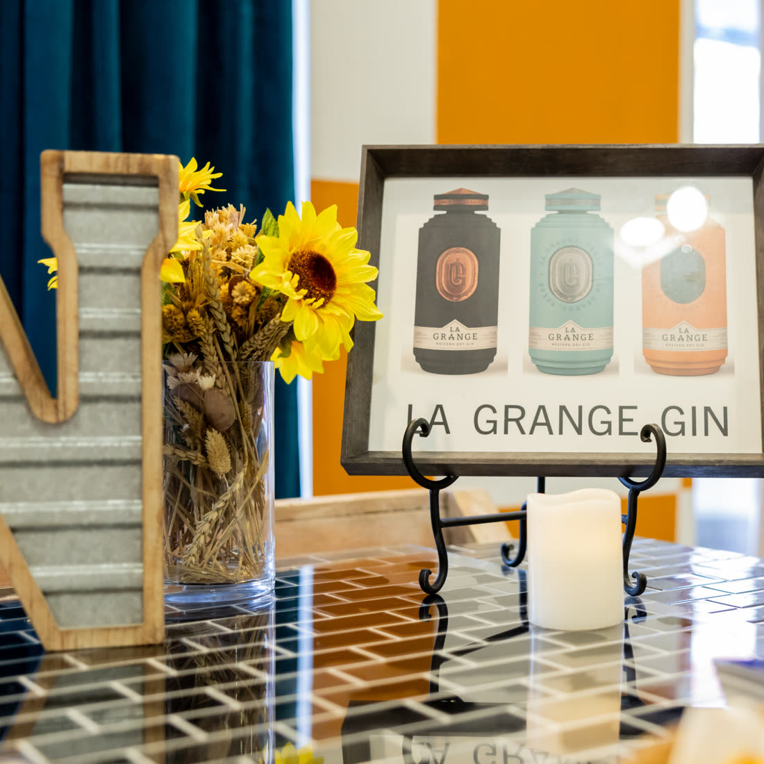 Framed La Grange Gin poster on a tiled tasting table with sunflowers